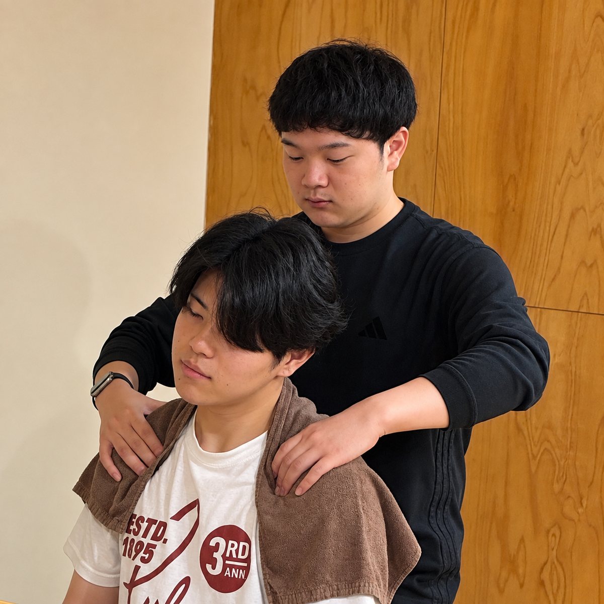 とんとん整骨院の練習会で、椅子に座ったスタッフの肩まわりに手を当てて施術の練習をするスタッフ
