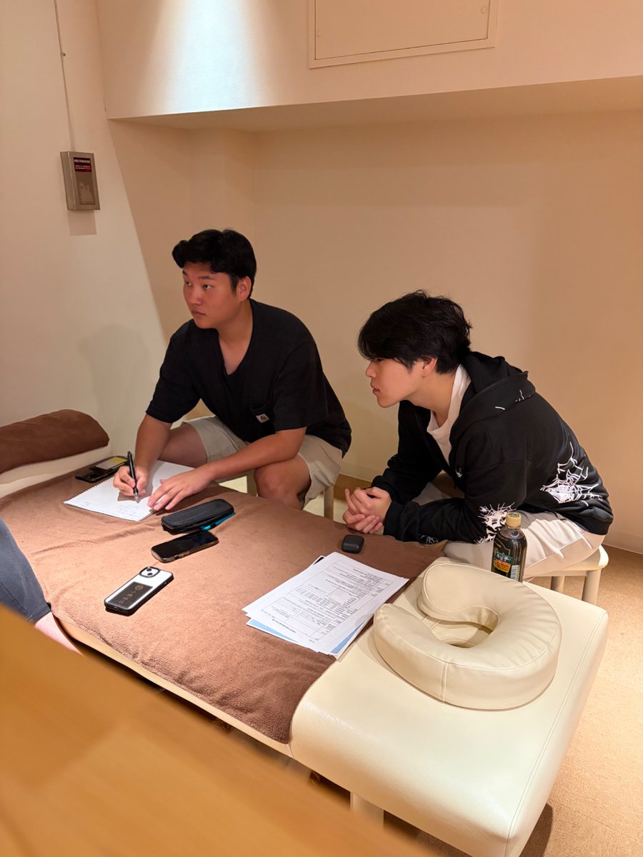 とんとん整骨院の練習会で、ベッドの脇に座ってノートに書き込みながら関連疾患の病態把握ロープレをするスタッフ2人