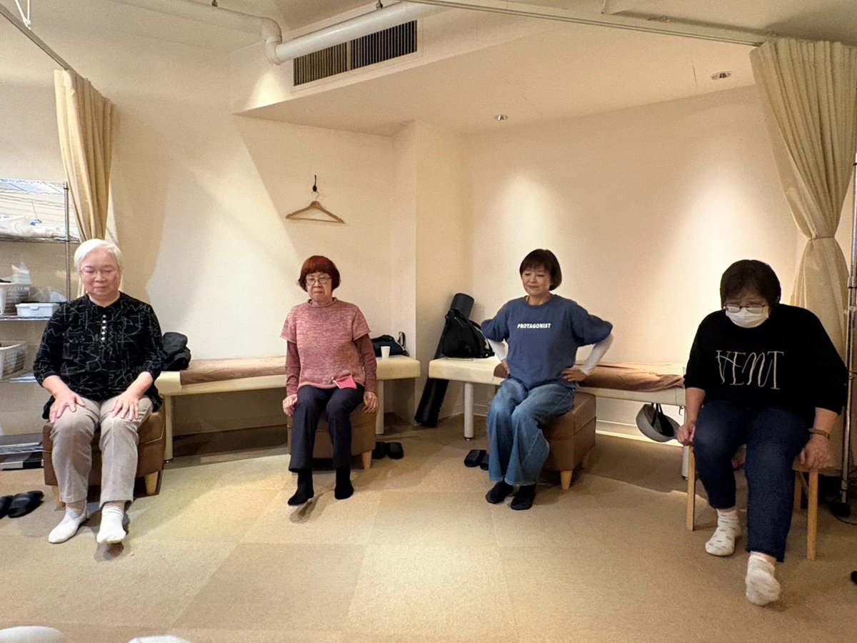 体操教室で、椅子に座った4名の参加者が横に並び、腰に手を当てて体操している様子